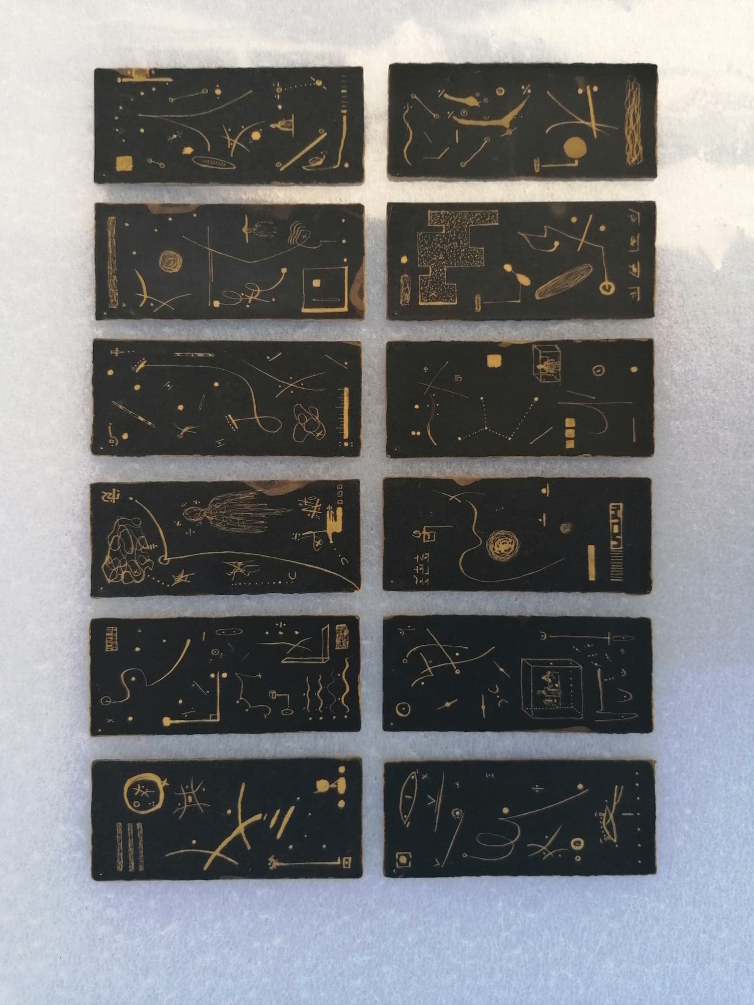 Twelve black cards laid out in two columns of six on a white sheet, each drawn in pale gold with constellations, tools, seed heads, small framed pictures and rows of marks