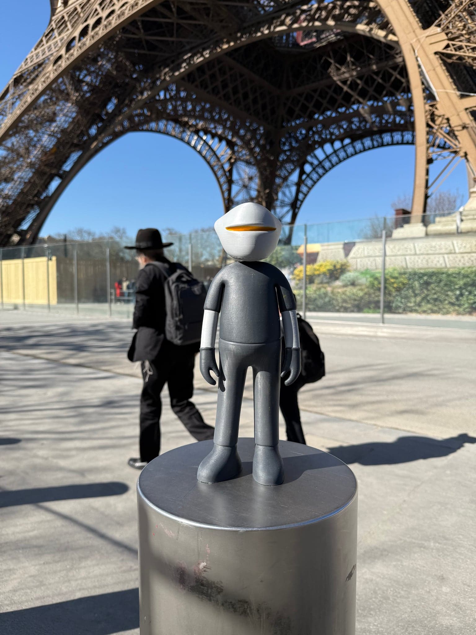 Mr. B on a bollard beneath the Eiffel Tower while a passer-by walks by in the background