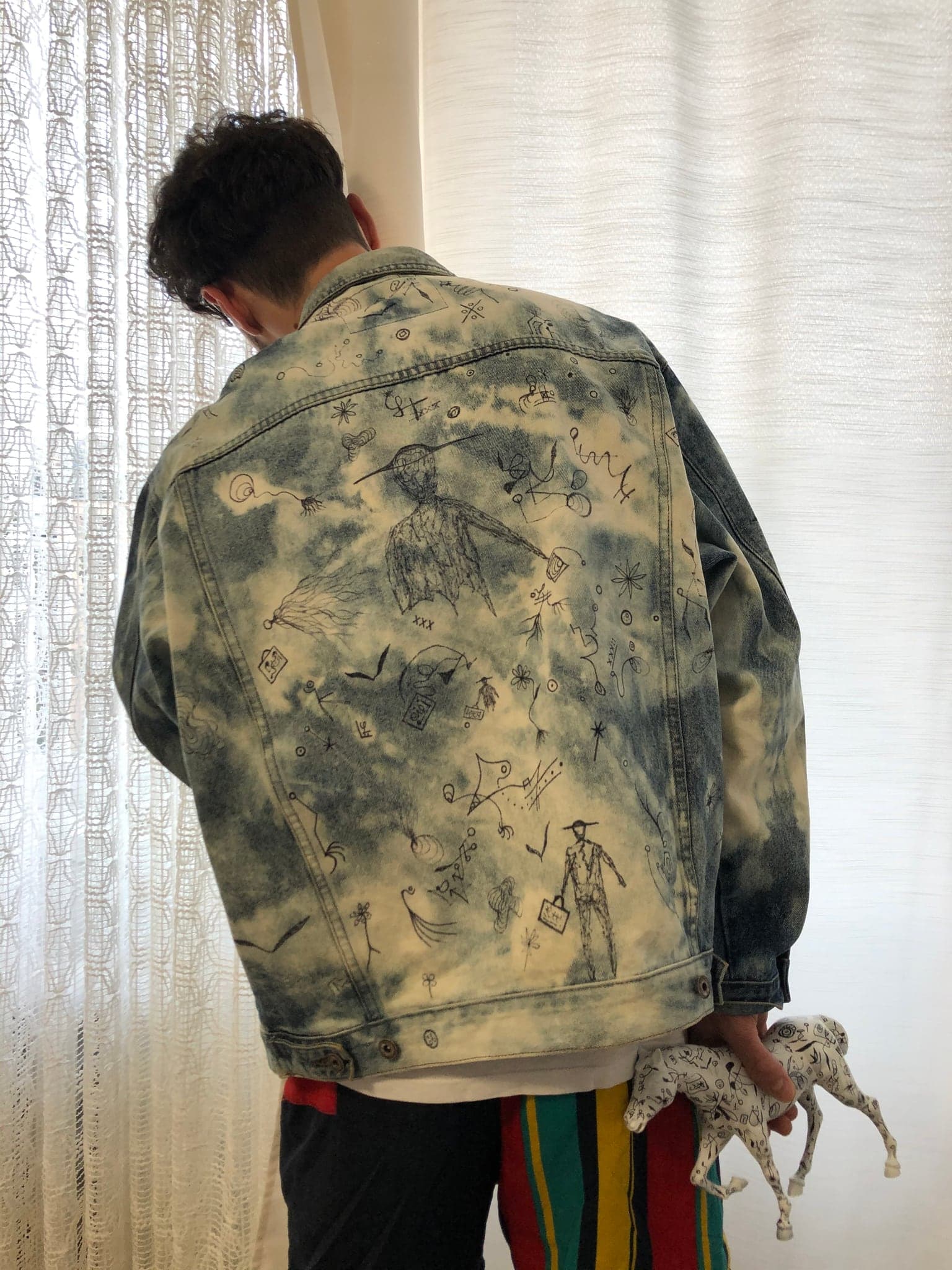 The back of a pale bleached blue denim jacket, worn, covered in fine dark line drawings scattered across the shoulders and body
