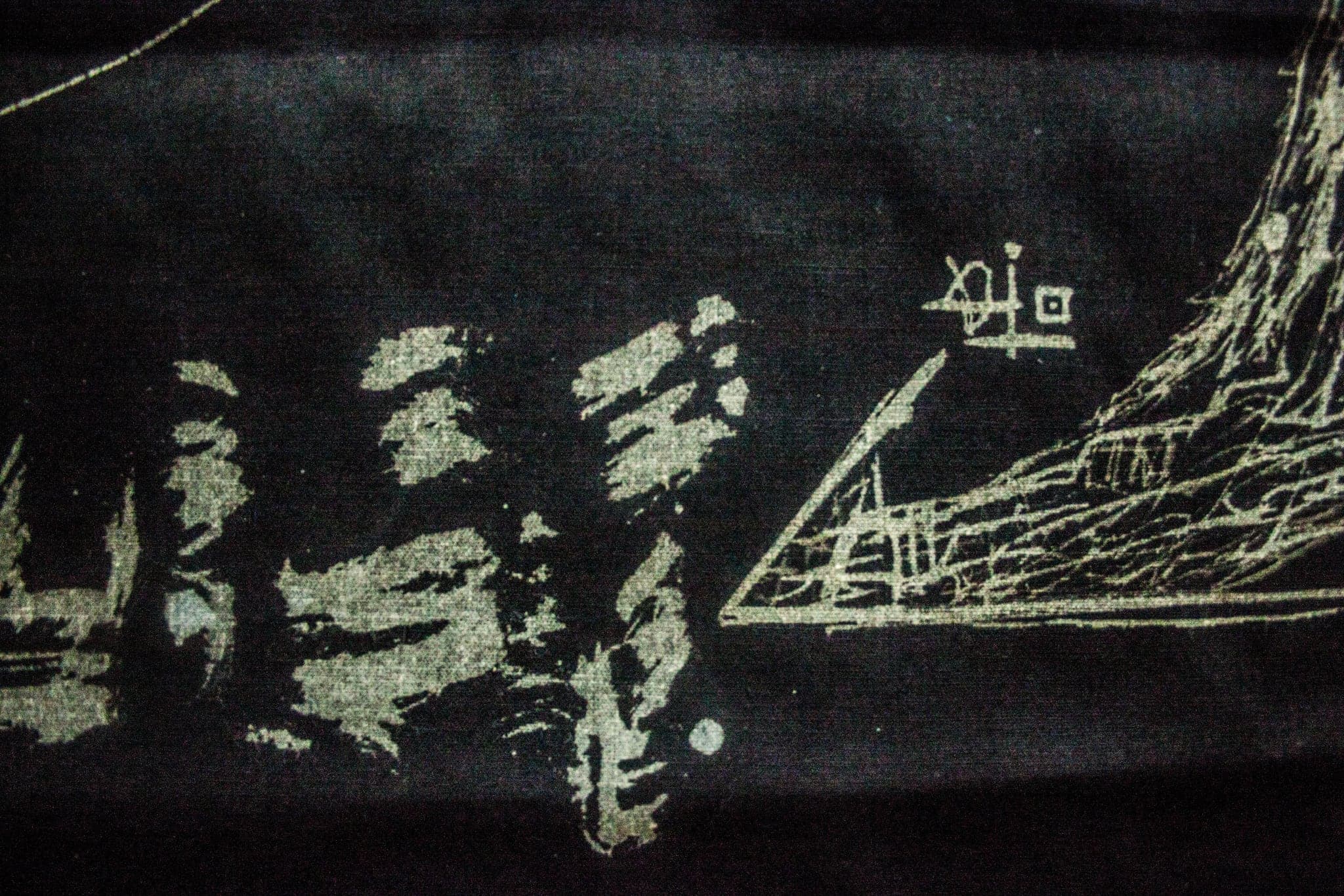 Close view of bleach on black cloth: torn pale blocks at the left, a drawn triangular structure at the right, a small glyph above it. A frayed thread runs loose across the lower edge.
