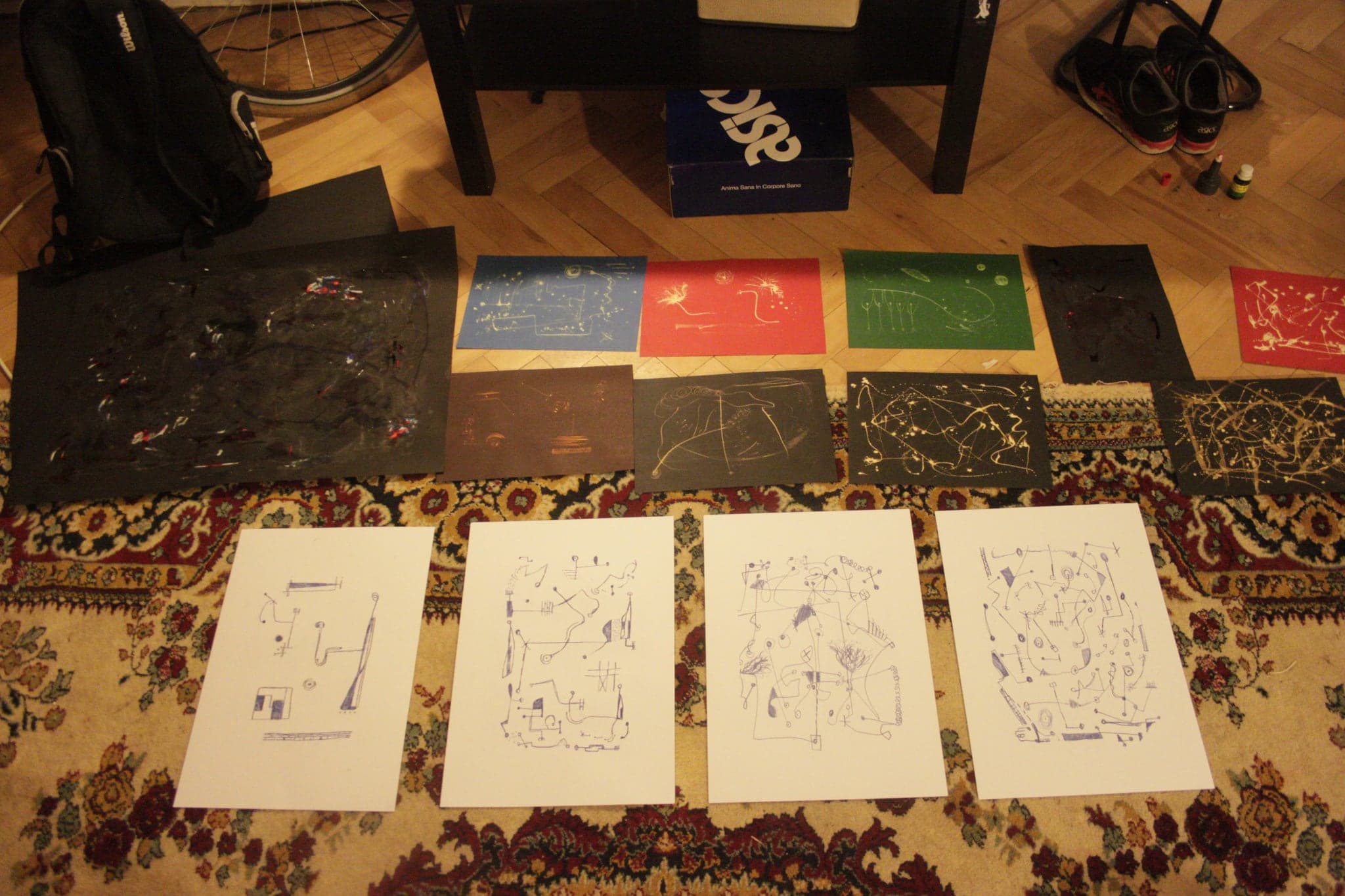 Works laid out on a patterned carpet: a row of dark and coloured sheets along the top, four white sheets with blue ink drawings below them