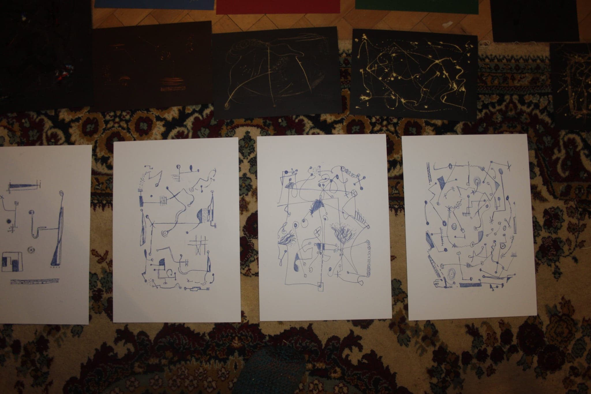 Black sheets marked in gold arranged along the top of a carpet with four blue ink drawings on white paper laid out beneath them