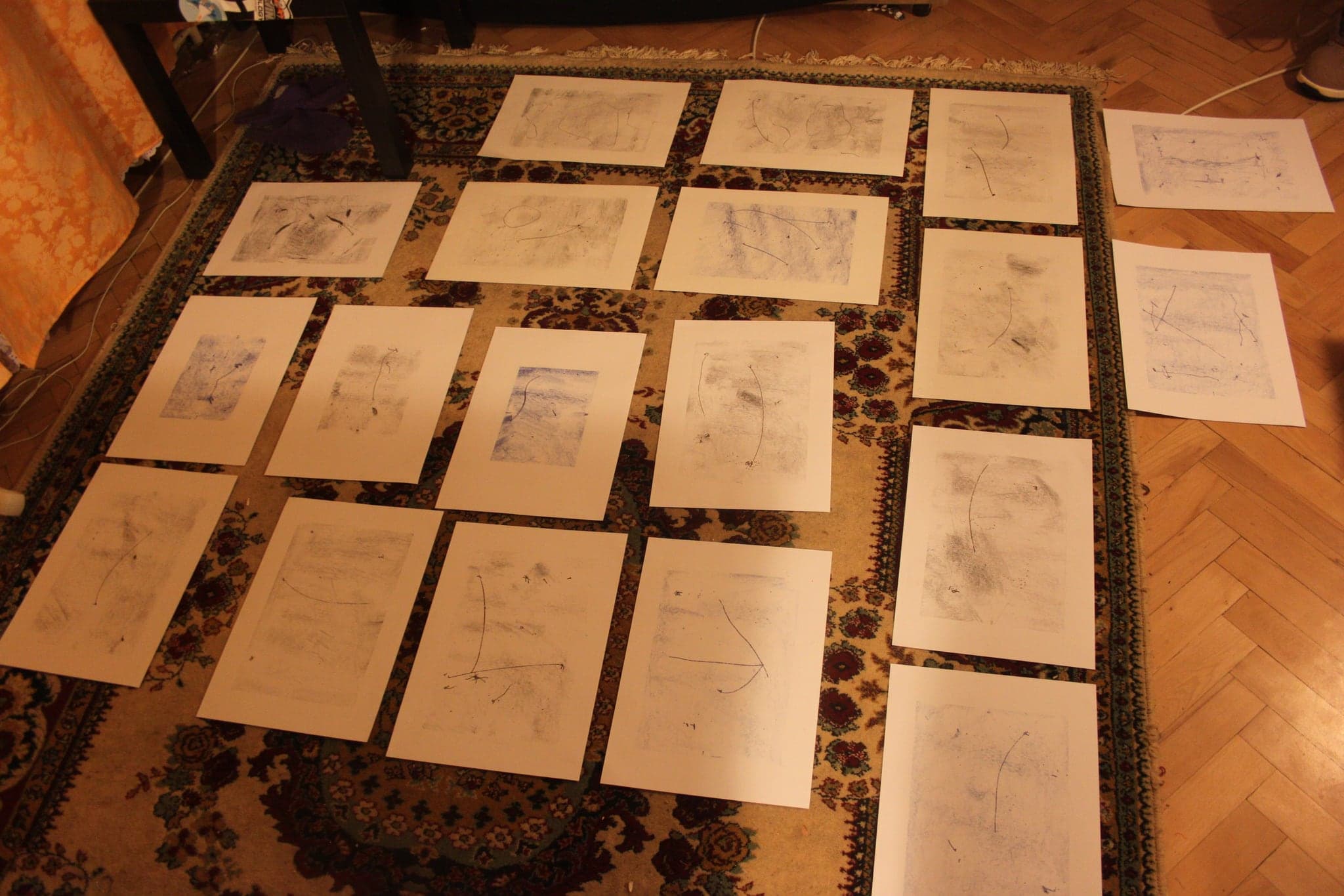 More than twenty pale drawings laid in rows across a carpet and parquet floor under warm lamplight, seen from above