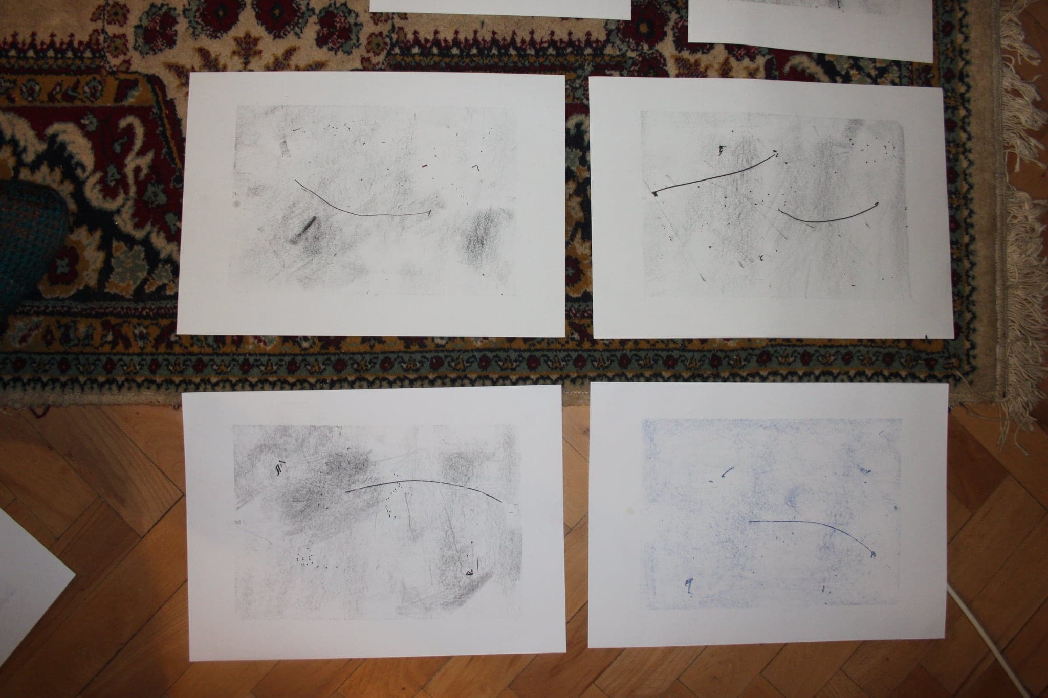 Four large white sheets on a carpet, each with sparse graphite marks, a smudged mass and a single long drawn line