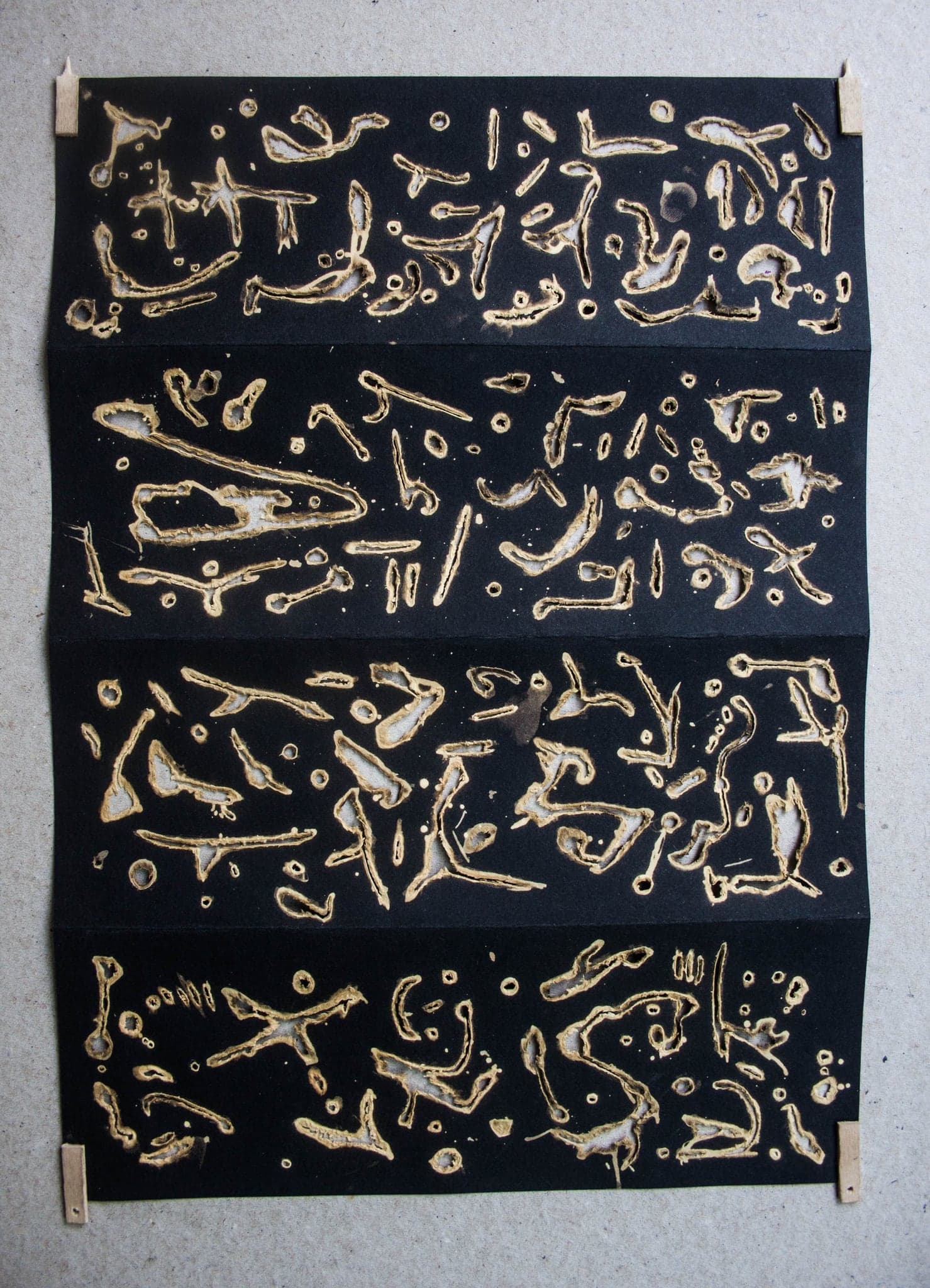 A sheet of black paper pinned at the corners with small wooden pegs, holding four horizontal bands of invented letter-shapes burned pale gold through the paper.