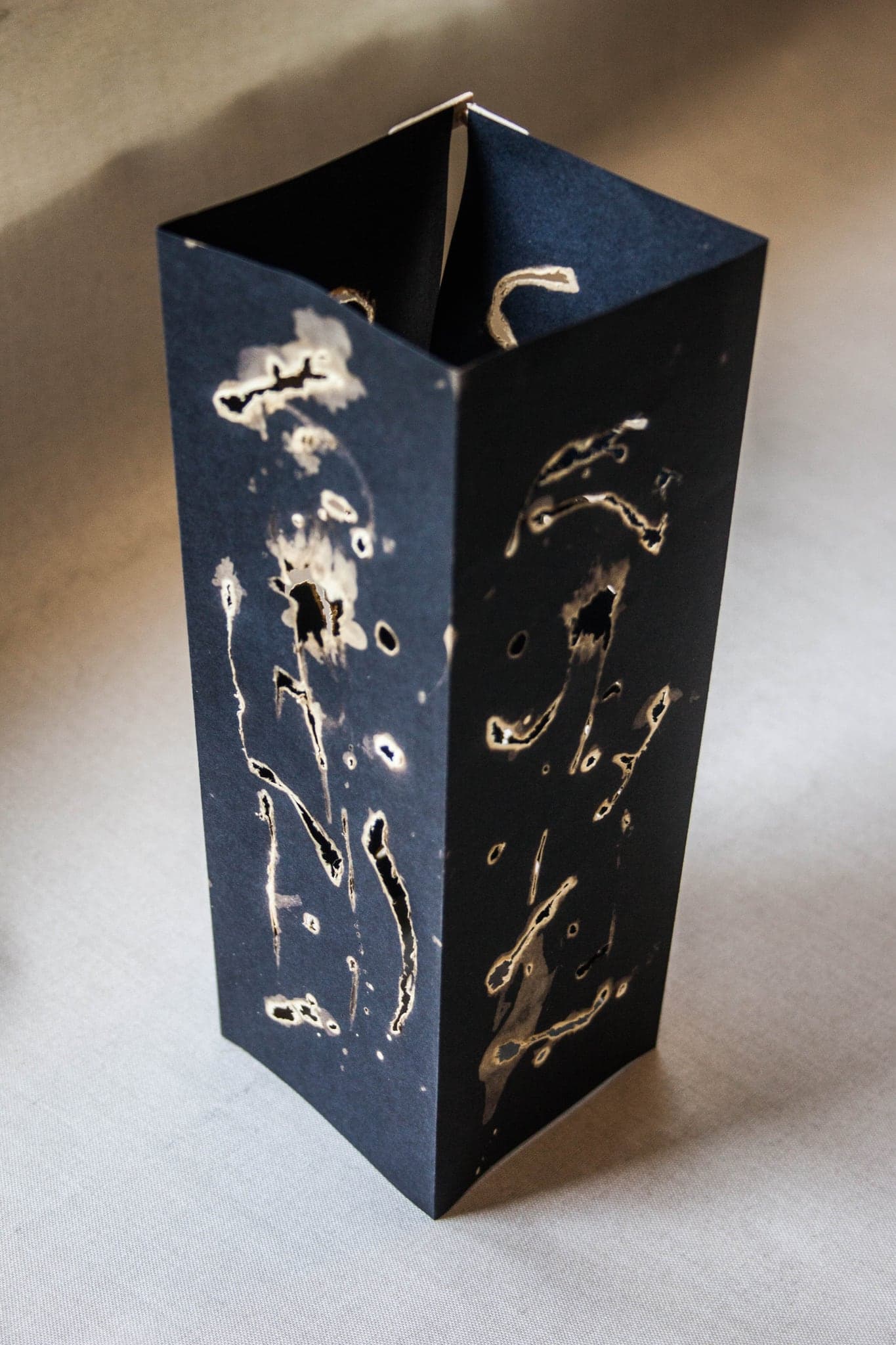 A tall square-section column folded from blue-black paper, standing on a pale cloth, its faces marked with pale eaten shapes.