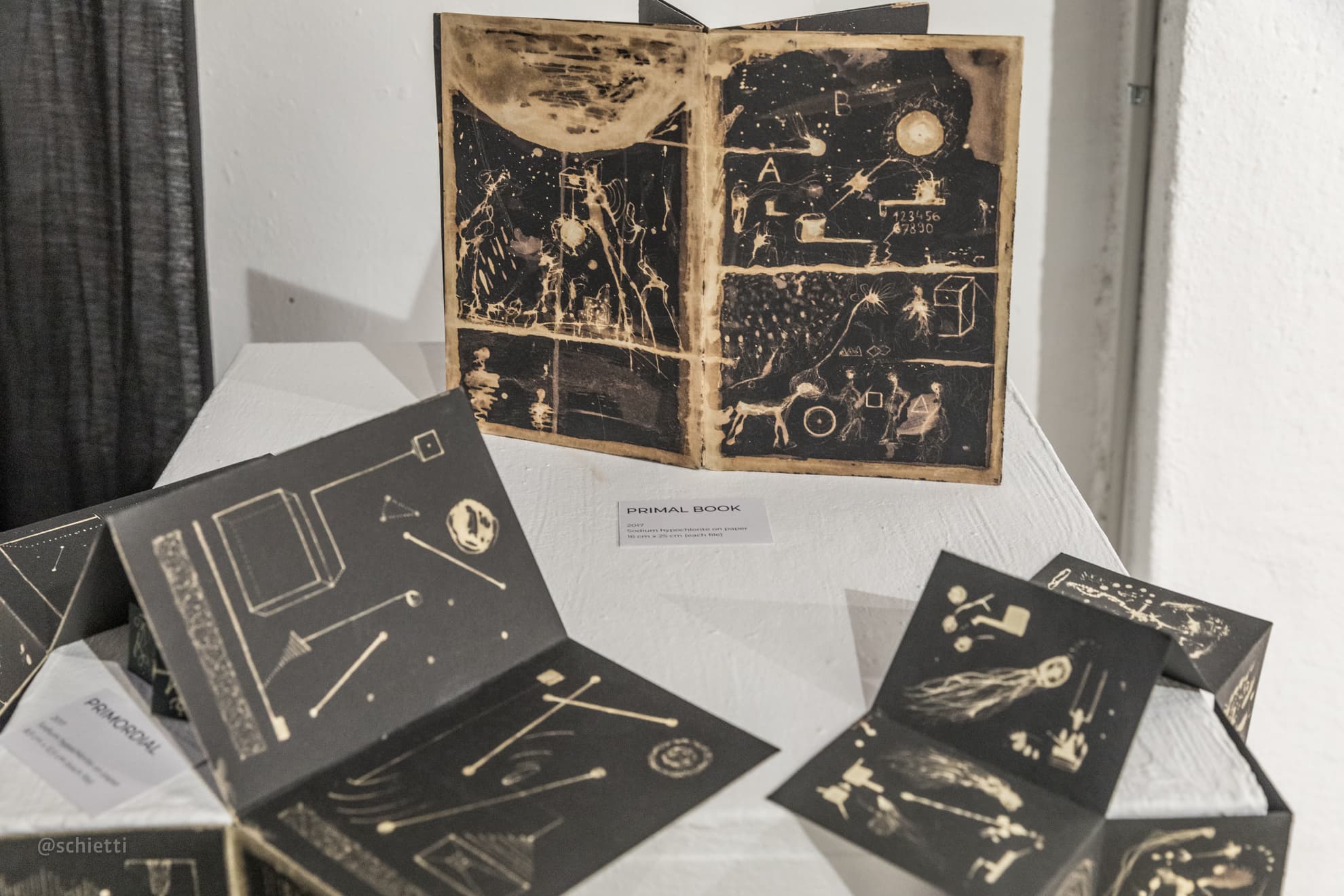 Several of the folded books standing open on a white plinth in a gallery, one propped upright behind them showing two pages of pale drawing, a small printed label beside them