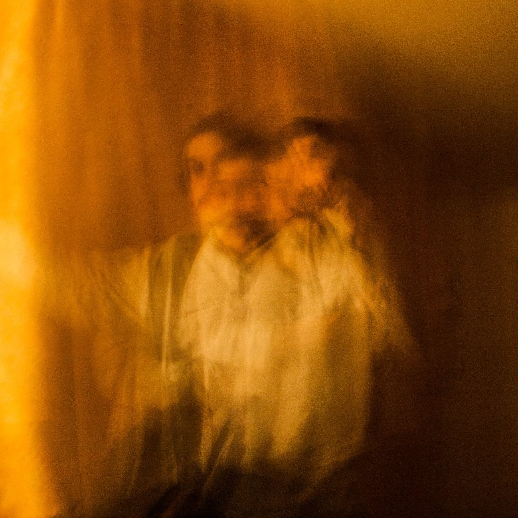 A warm orange interior in which a figure doubles and blurs across a long exposure, face and shoulders dissolving into the wall behind.