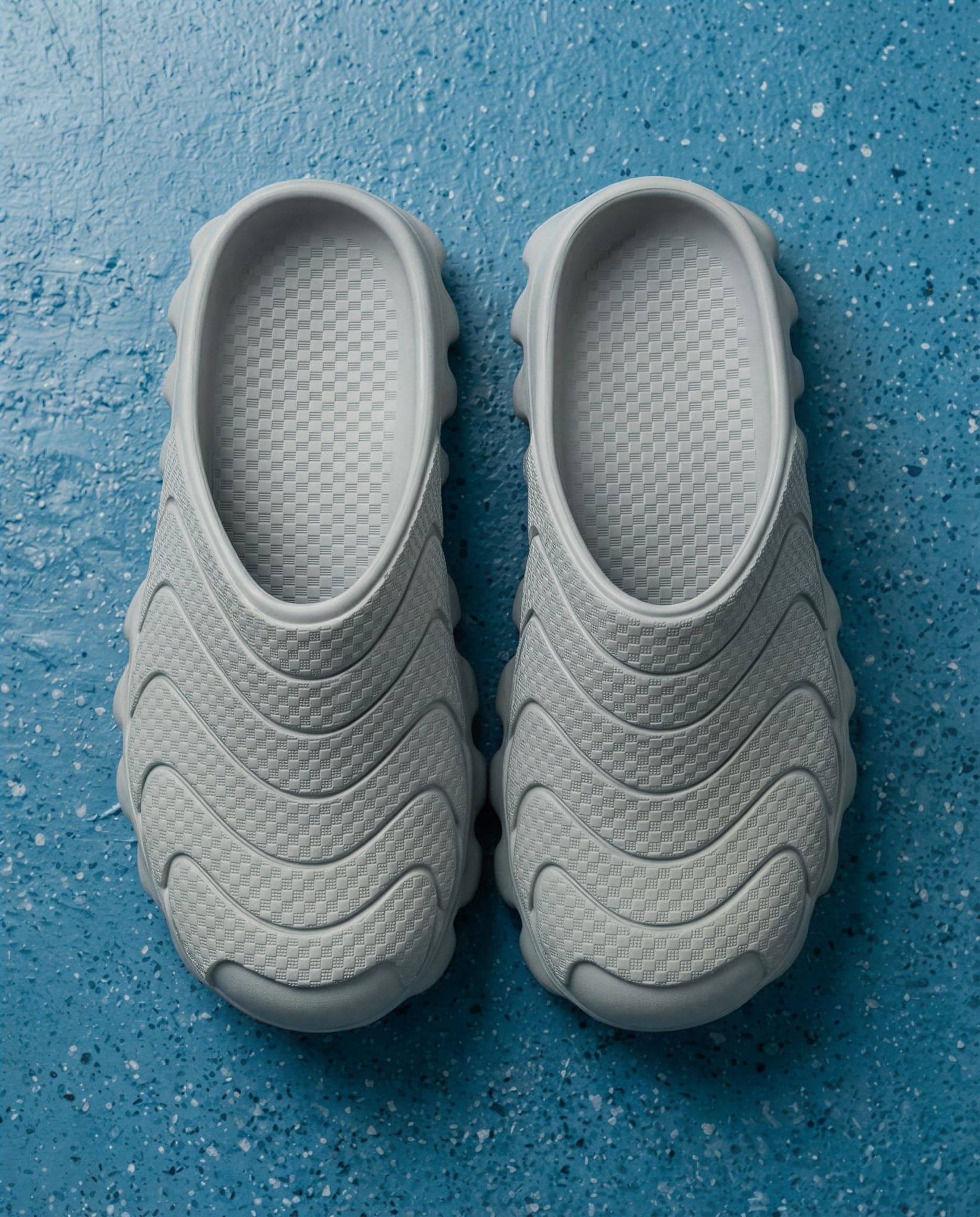 Top-down view of a pair of pale grey backless mules on a blue speckled floor, uppers ribbed with woven wave bands.
