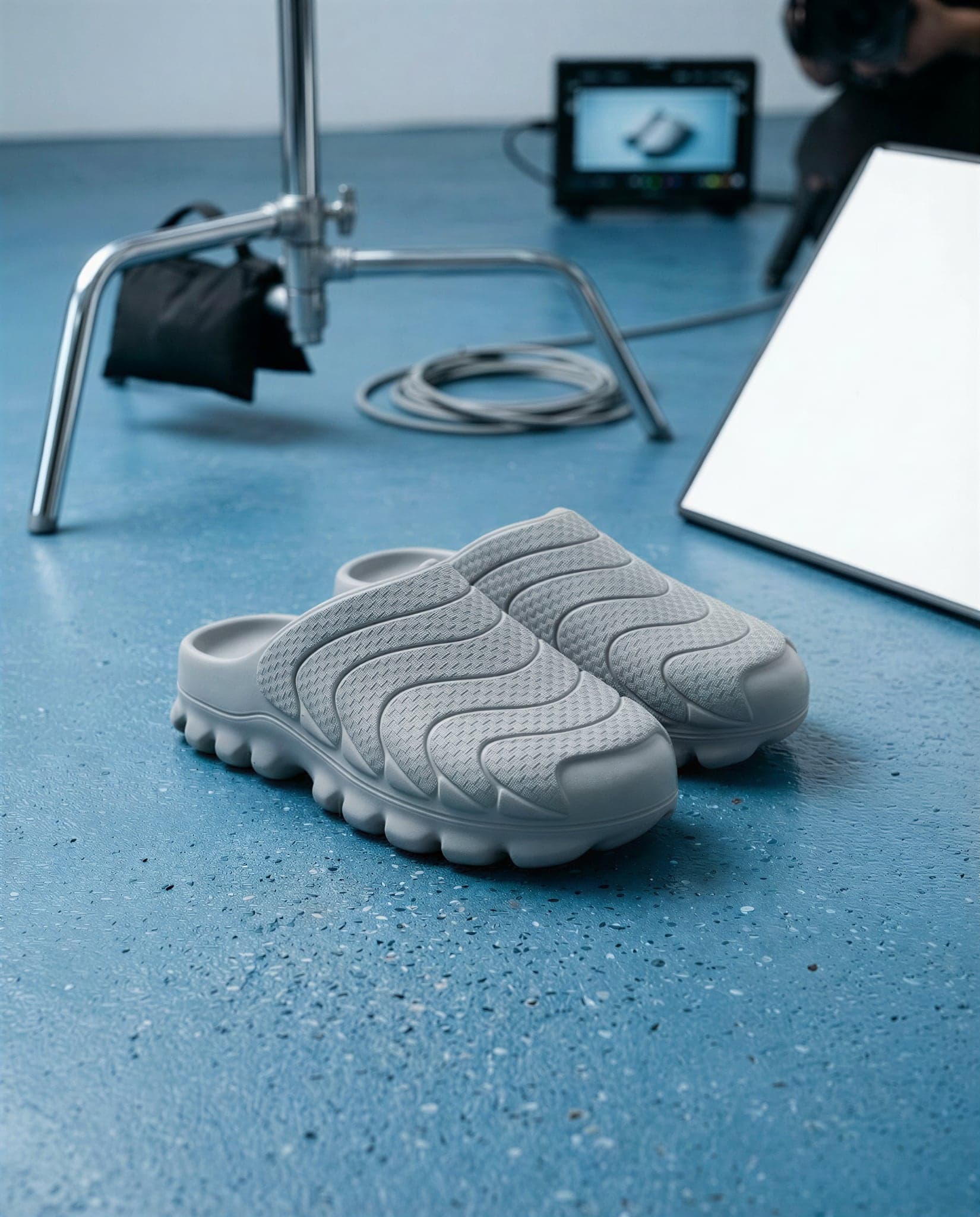 The pair of grey mules on a blue studio floor with a light stand, cable coil and monitor behind them.