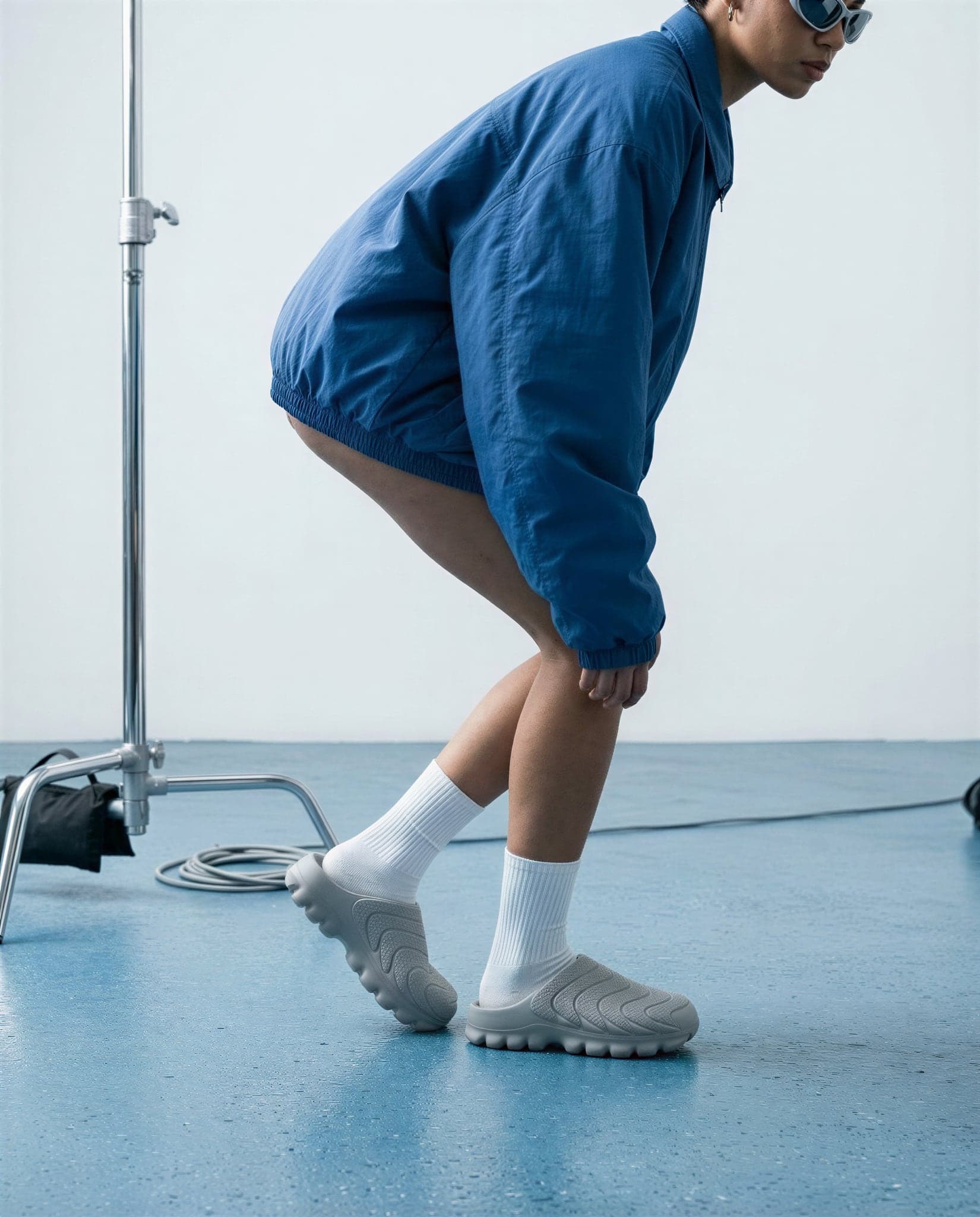 A person in a blue jacket crouching, wearing the grey mules with white crew socks on a blue floor.
