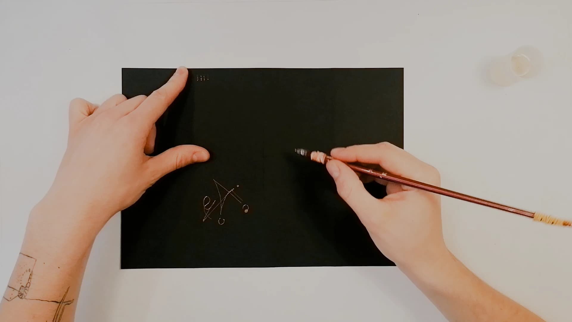 A nearly blank black sheet on a white table. One hand steadies the paper, the other draws with a wooden nib holder. Two small pale marks have appeared.