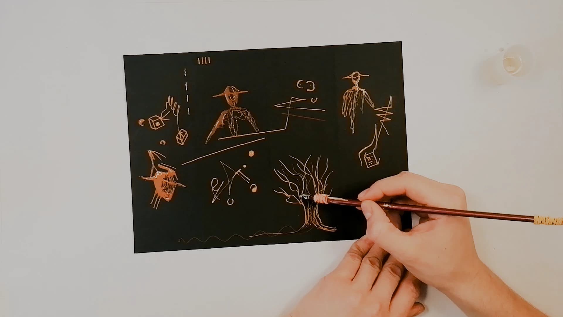 The sheet about two thirds filled. A bird-headed figure in a wide hat, a standing man, a horned head and a tree, all in copper bleach lines on black. A hand works at the tree with a nib.