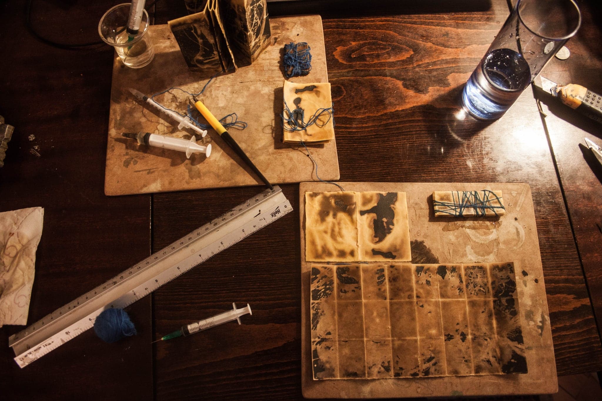 The working table under a low lamp: a ruler, syringes, blue thread, and treated cards laid out on boards.