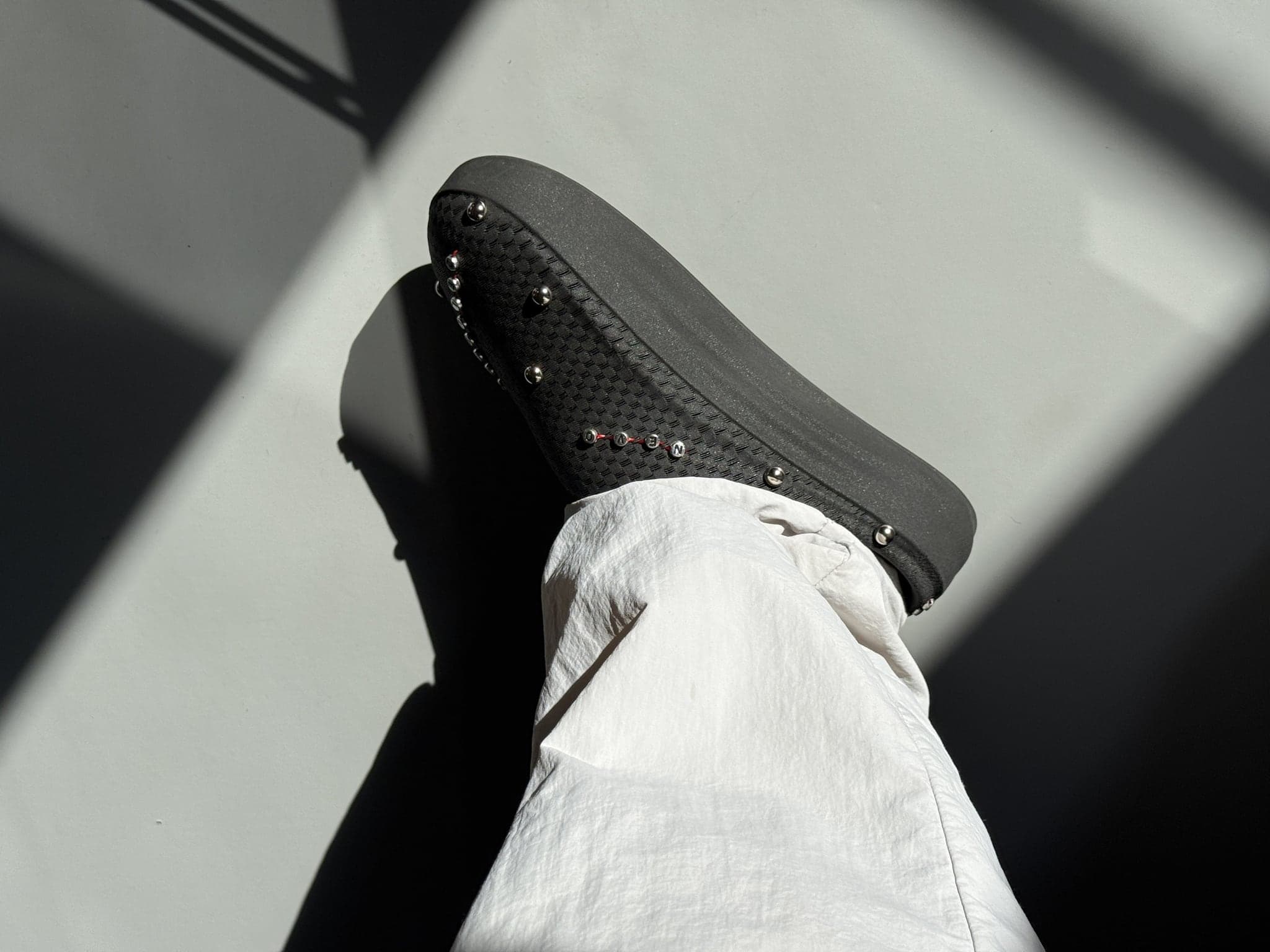 The customised black NEVO worn on the foot in hard sunlight with white trousers, the row of metal studs catching the light along the side