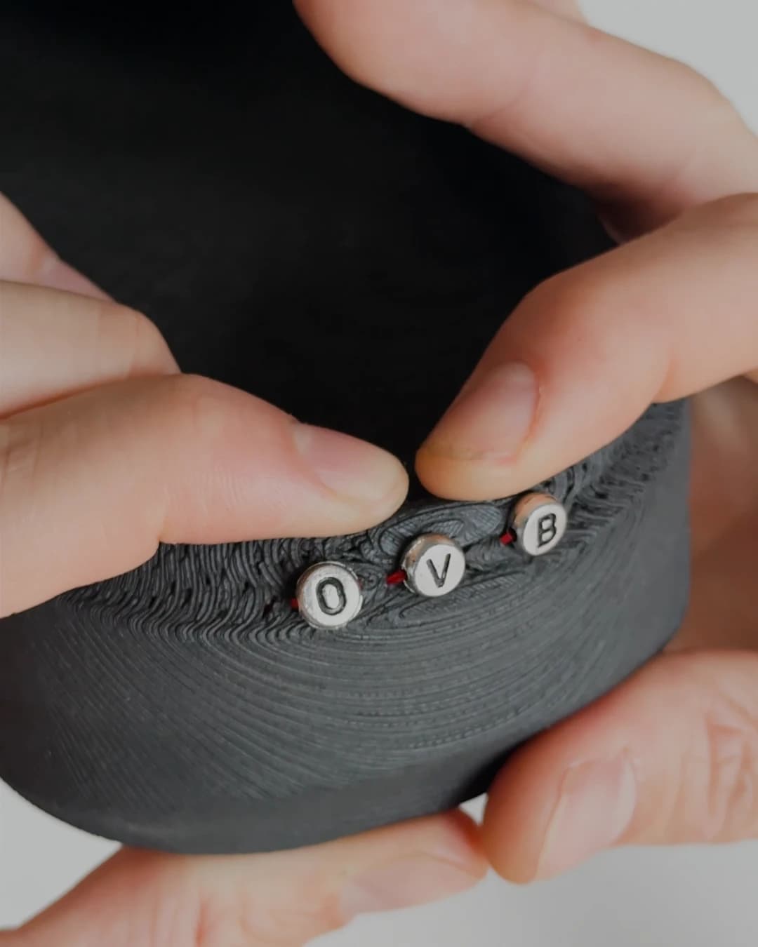 The lettered beads held between fingers with the red thread running away from them across the black surface