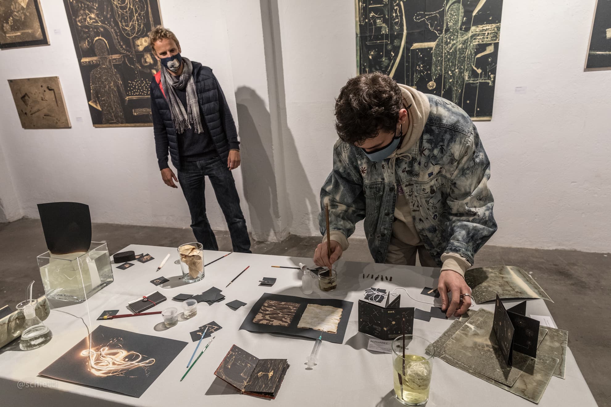 The artist bent over a white table in the gallery, working on a black card with a brush, while a visitor stands watching; bleached works hang on the wall behind