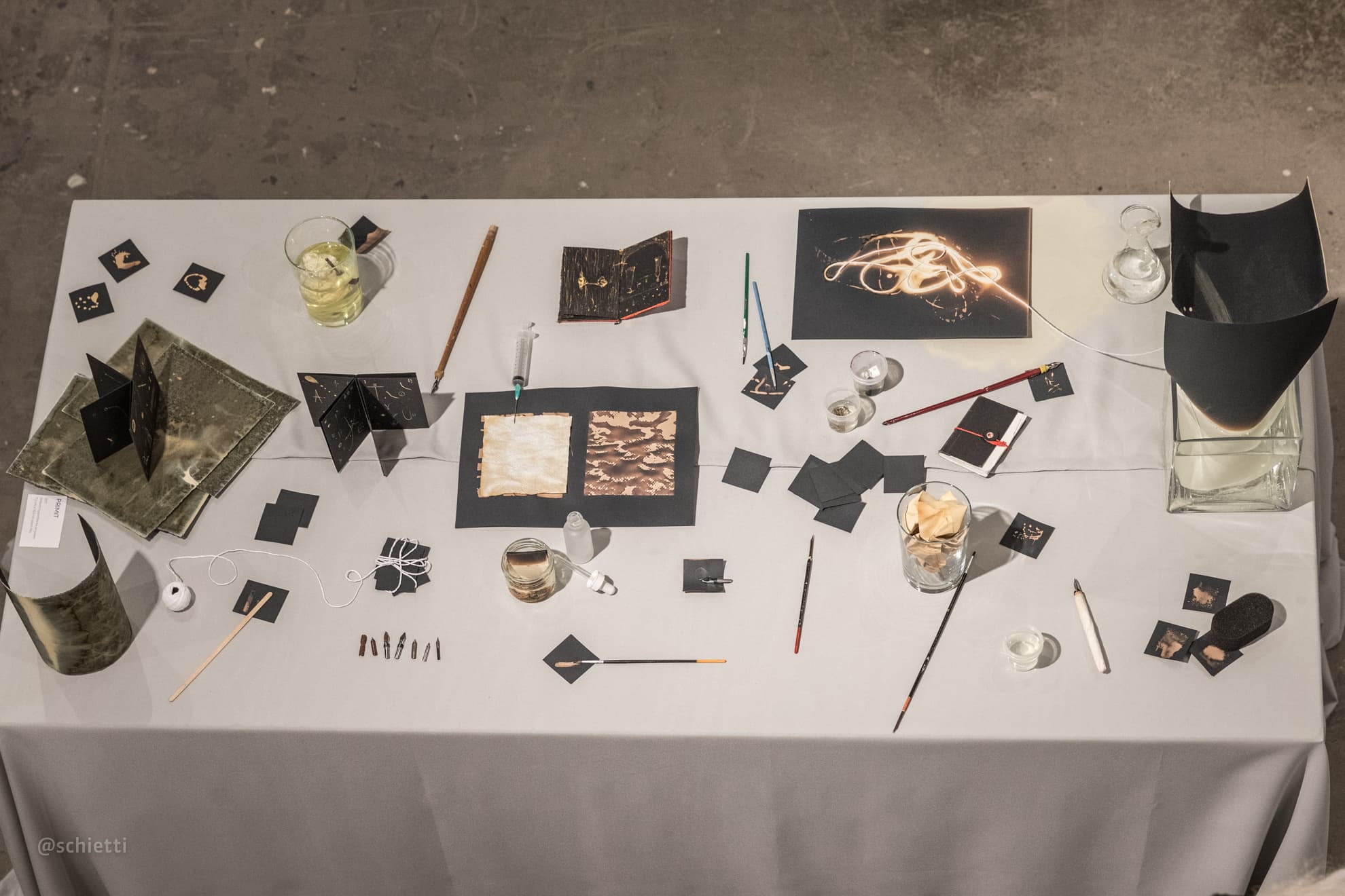 The whole white table seen from above, covered in black cards, folded books, glasses and jars of bleach, brushes and small tools