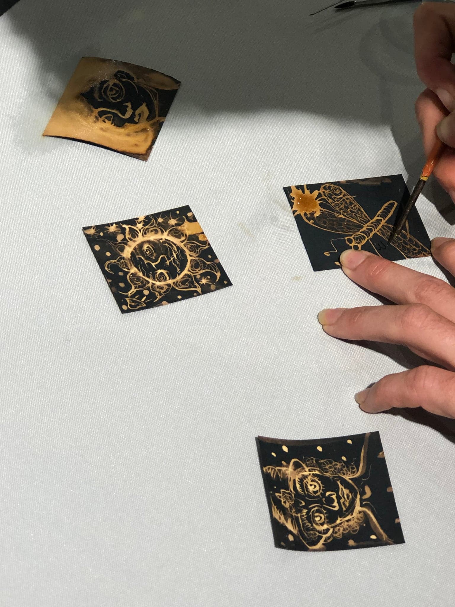 A close view of two hands drawing on a small black card with a nib, three finished cards with gold marks lying beside it