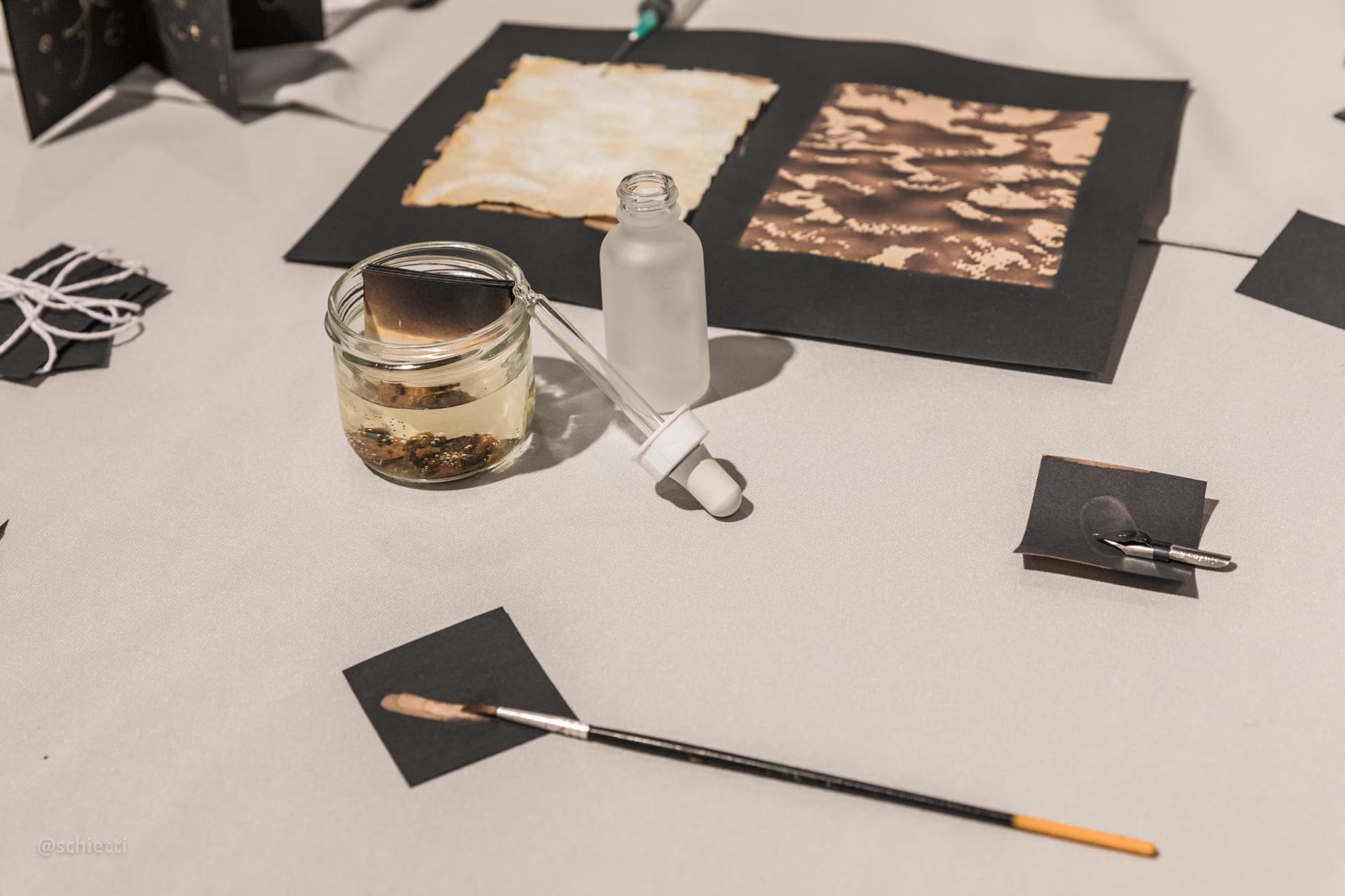 A jar of bleach gone brown, a dropper bottle and a brush lying on the white cloth beside two mounted sheets