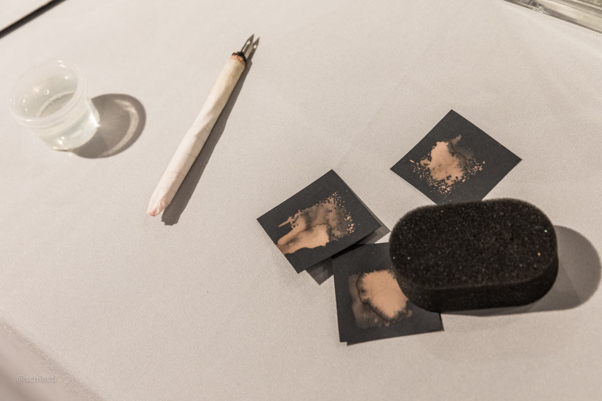 A rolled paper tool, a glass of water, small black cards and a black sponge on the white cloth