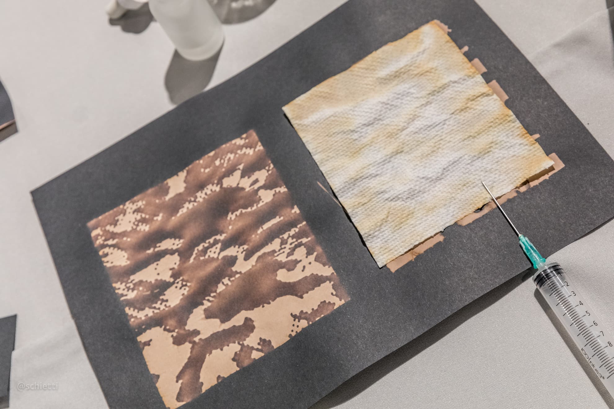 Two sheets mounted on a black card — one copper and mottled, one cream and streaked — with a syringe lying across the corner
