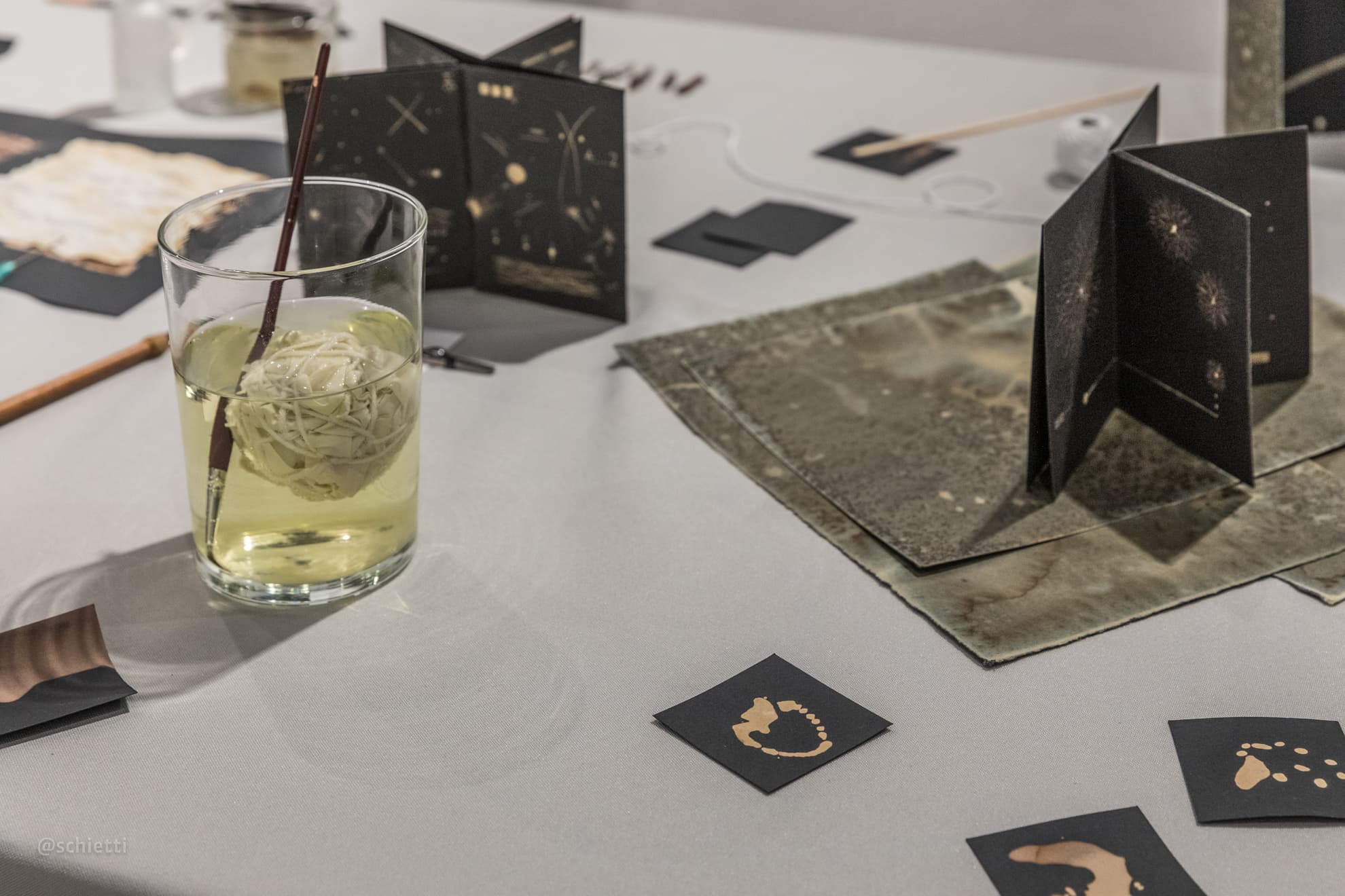A glass of bleach with a brush standing in it, black cards and small folded books spread across the white table