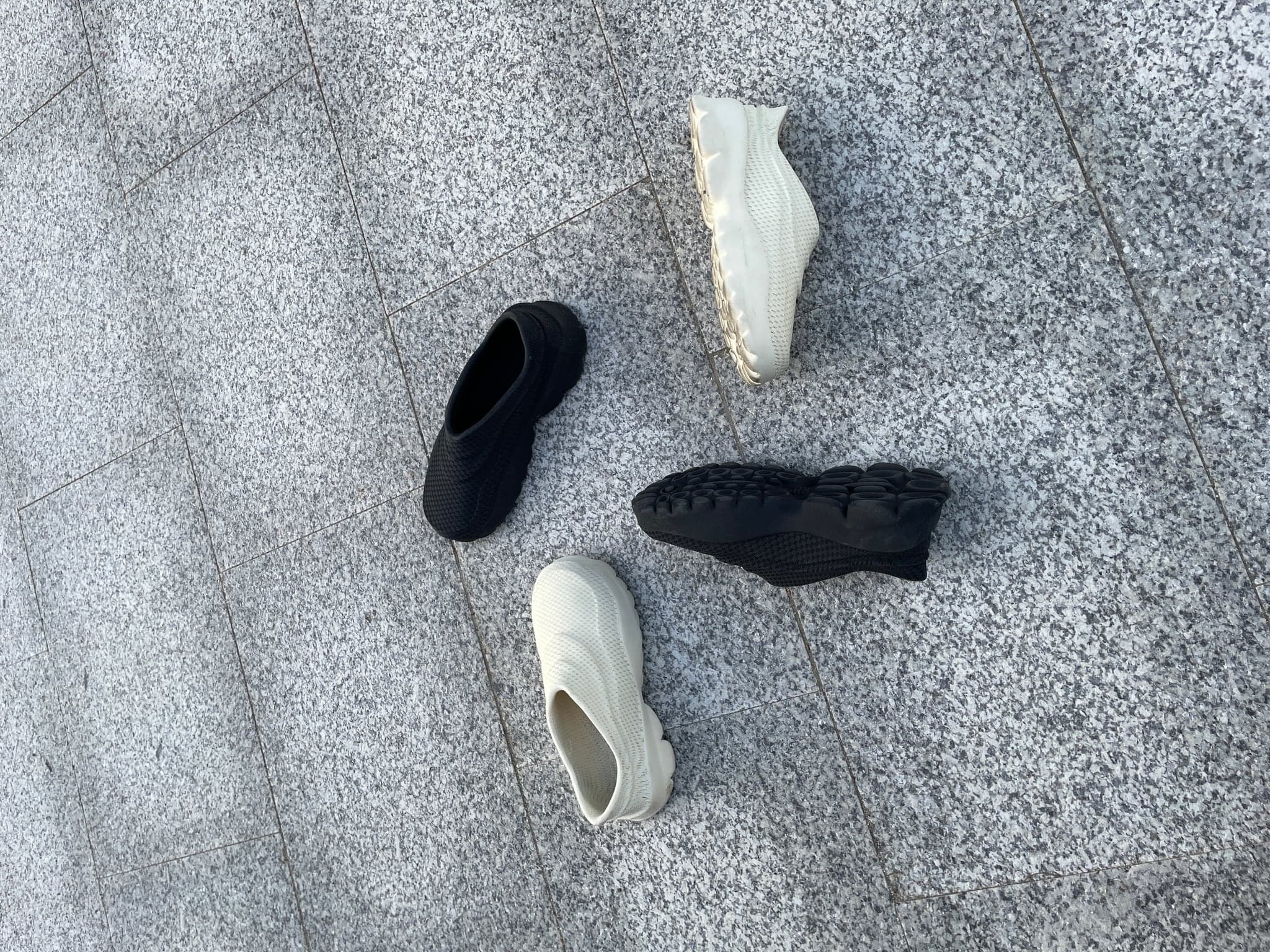 Four shoes arranged on pale granite paving: two bone and two black, one turned over to show the oval-pod tread.