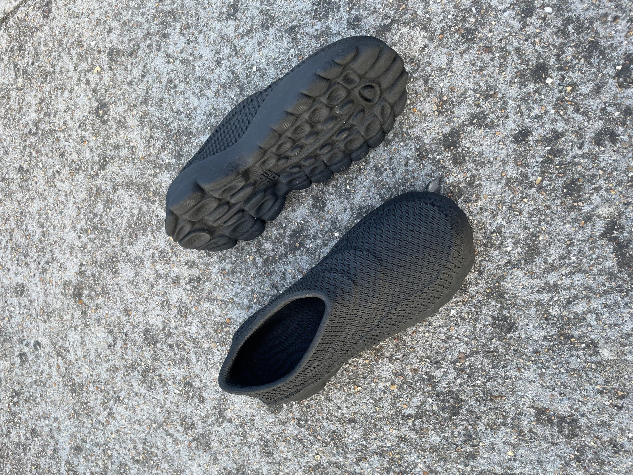 A black pair on rough concrete, one shoe upside down showing the pod tread, the other upright showing the knitted upper and open ankle.