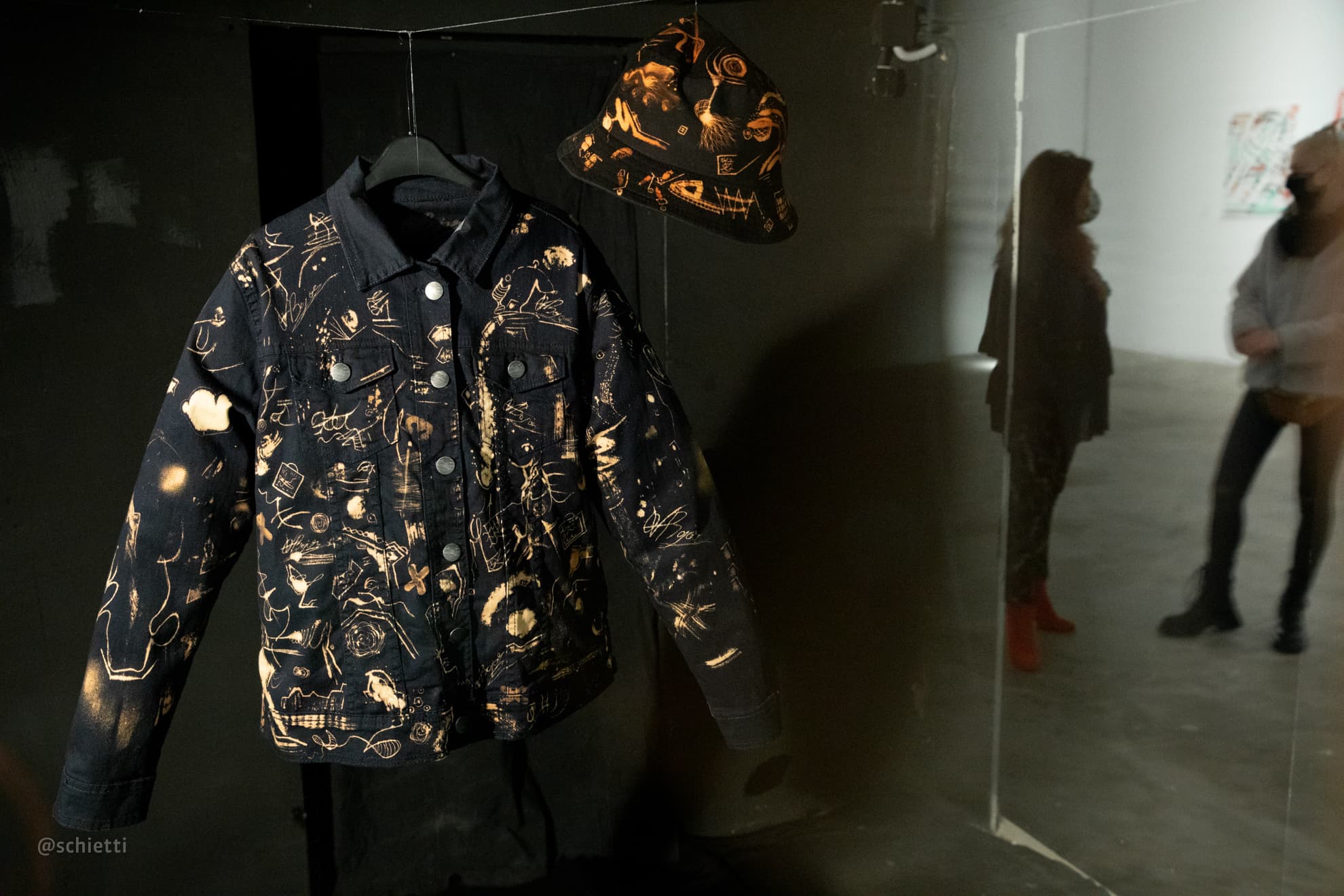 The jacket hanging in a dark gallery beside a gold painted hat, visitors in the lit room beyond