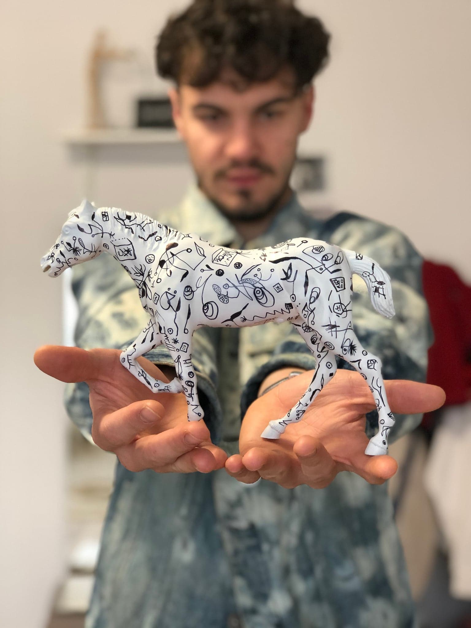 The paper horse standing on two open hands held out towards the camera, the artist behind it in a bleached denim jacket