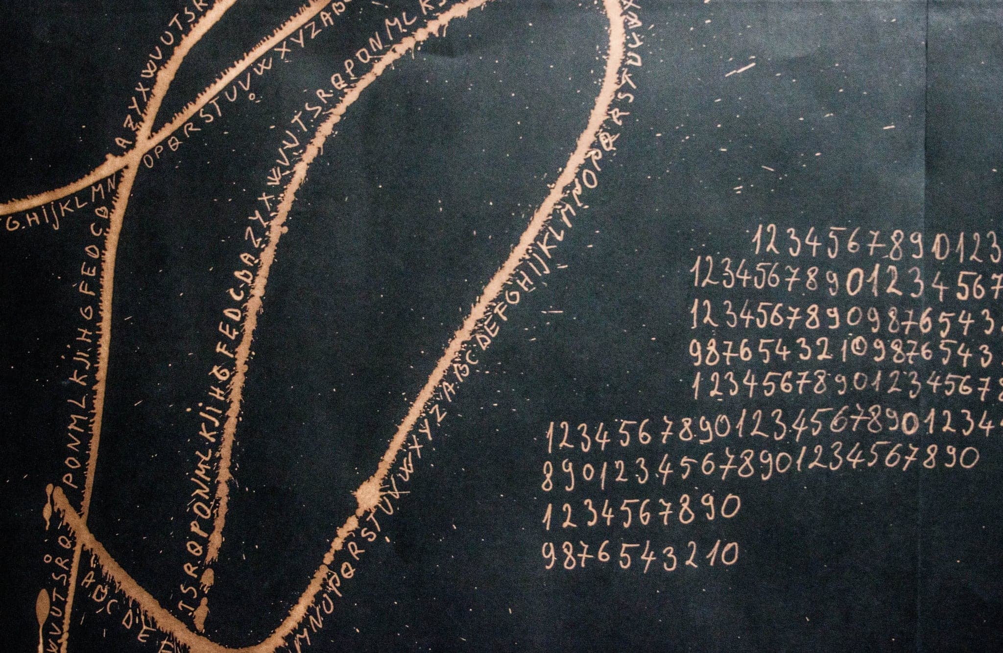 Detail of a black sheet: bleached lines with alphabets written along them in pale gold, and columns of handwritten digit sequences running down the right side.