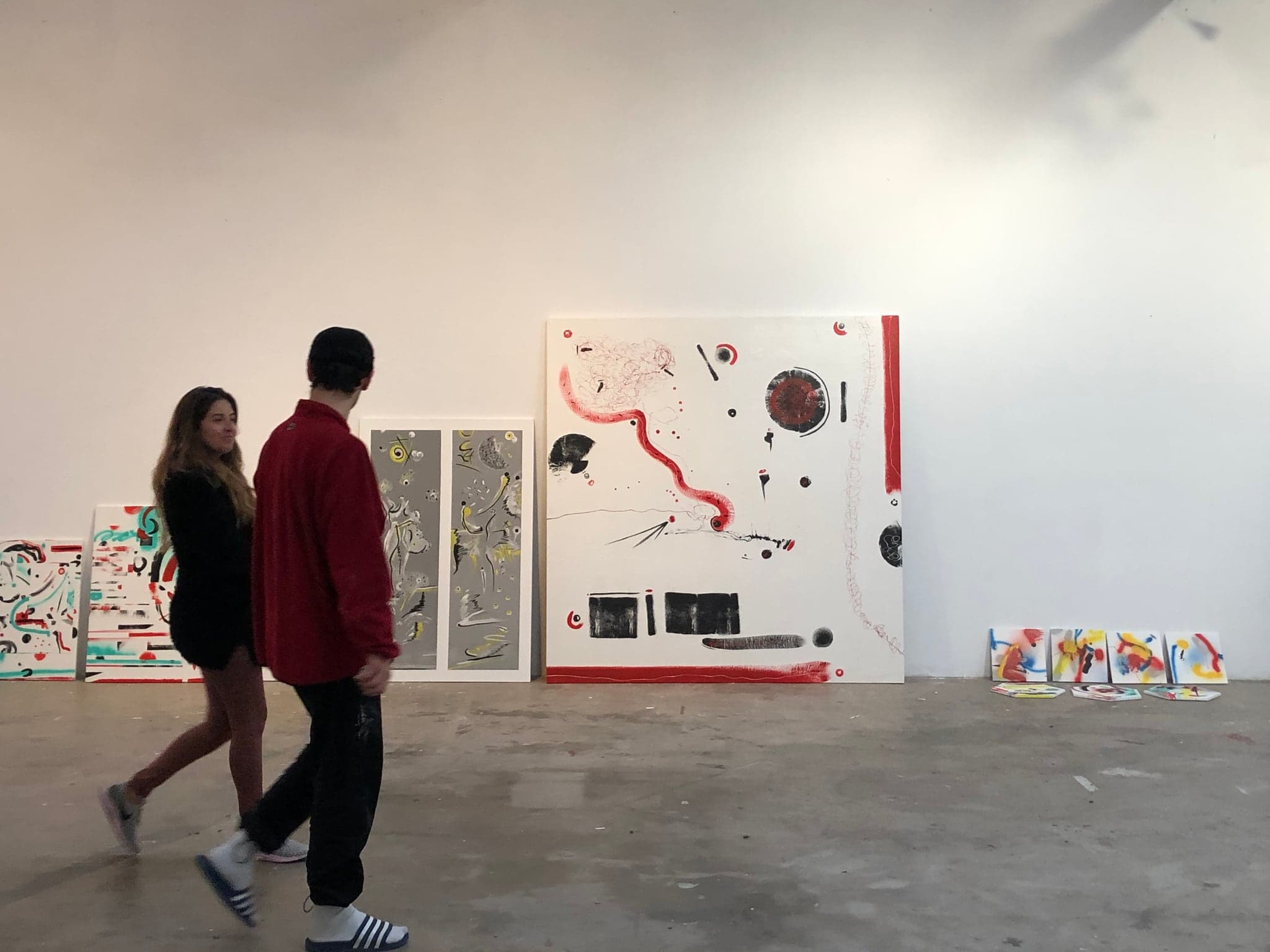 Several of the paintings leaning unhung against a gallery wall, the four small spray squares propped on the floor beside them, two people walking past