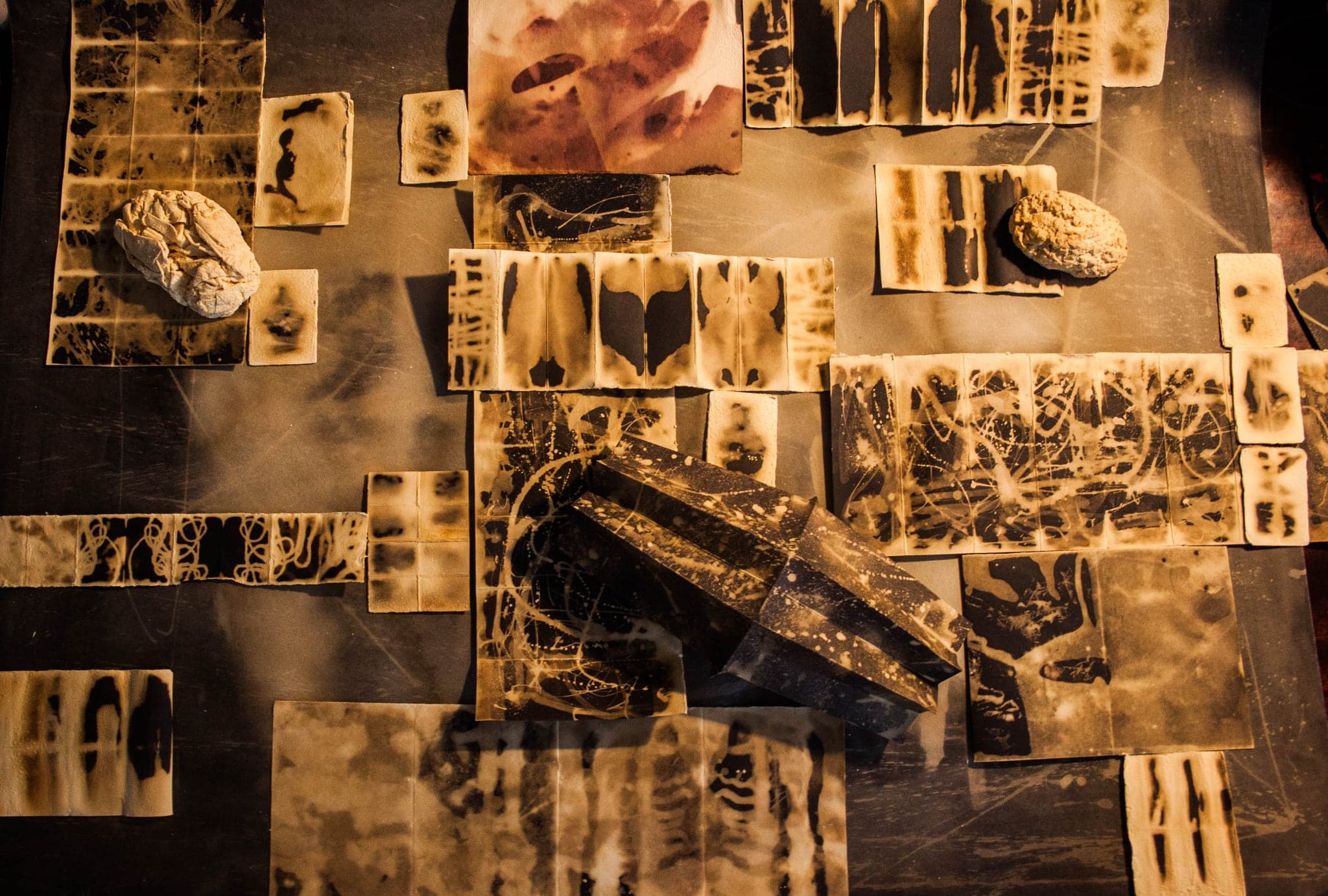 Overhead view of dozens of amber-lit wax tiles arranged on a dark worktable, black bars and blots inside each tile.