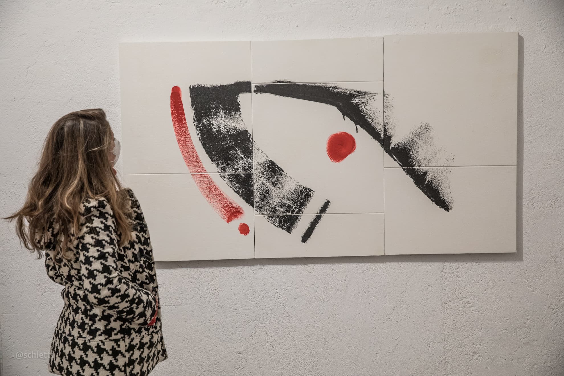 Six white panels hung as one horizontal field, crossed by a single sweeping black brush stroke, a red arc beneath it, a red disc in the middle and a short black slash; a visitor stands looking at it